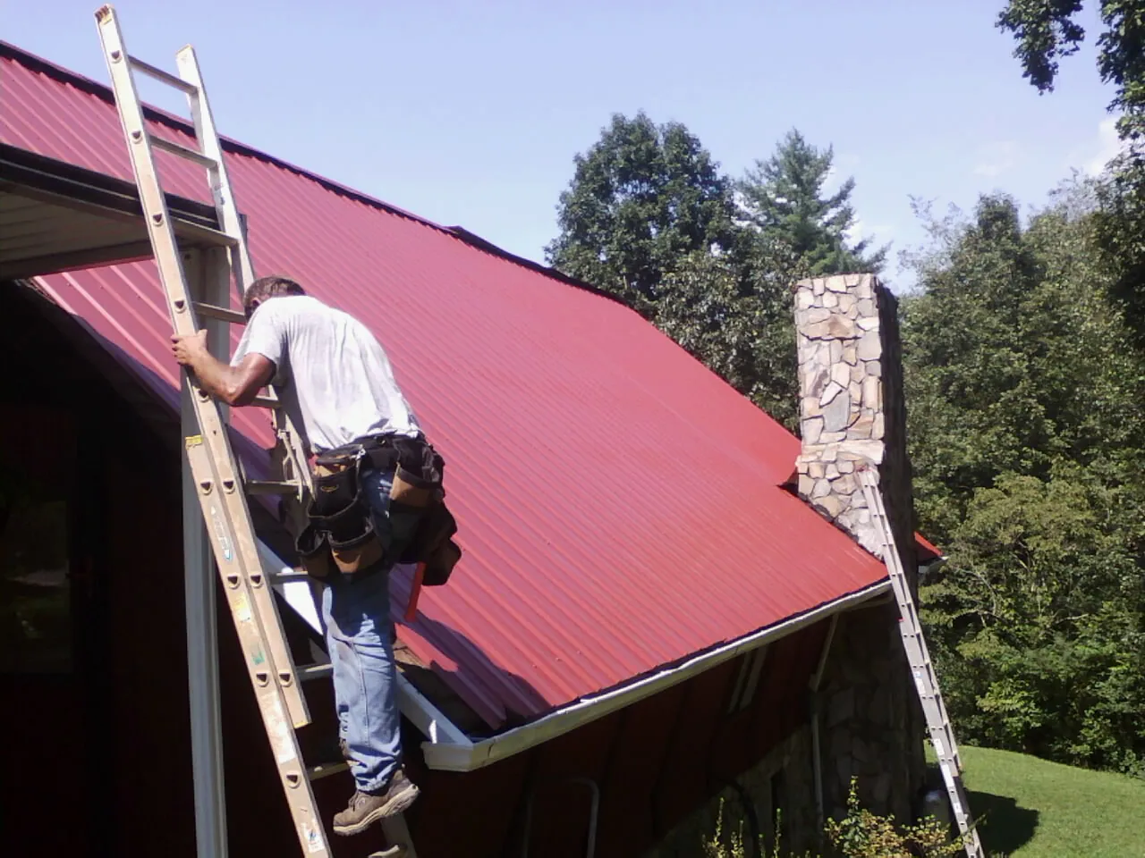 Roofer on a ladder beside a red metal roof and stone chimney — Soffit & Fascia Repair by Roofing Brothers in Gooding